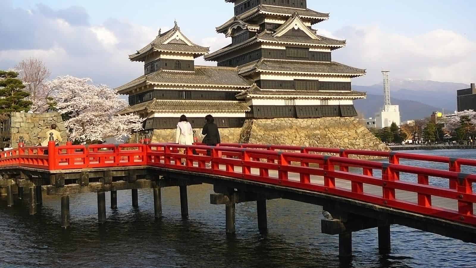 Two people walk across a red bridge toward a traditional Japanese castle surrounded by water and cherry blossoms&mdash;a picturesque scene that highlights one of the top Japan summer destinations.