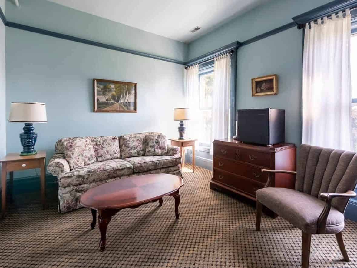 Bright living room with floral sofa, armchair, TV on dresser, coffee table, lamps, and large windows with curtains&mdash;perfect for relaxing after exploring Finger Lakes Wineries with Lodging nearby.