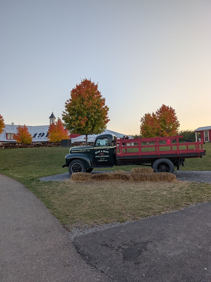 Old green farm truck parked on grass near autumn trees, hay bales, and a barn at sunset—perfect for a Finger Lakes fall road trip or weekend getaway.