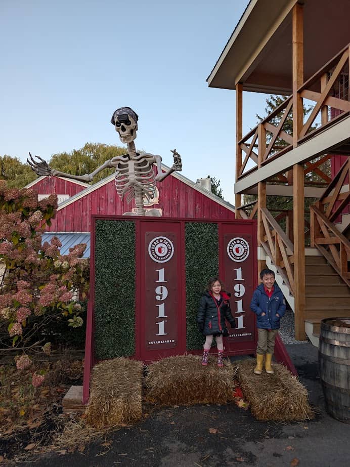 Two children stand on hay bales in front of 1911 signs with a large skeleton decoration above them—perfect for a Finger Lakes fall road trip.