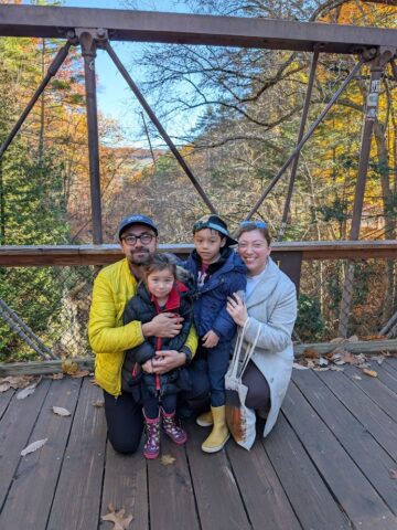 A family of four poses and smiles on a wooden bridge at Watkins Glen State Park, with vibrant fall trees in the background.