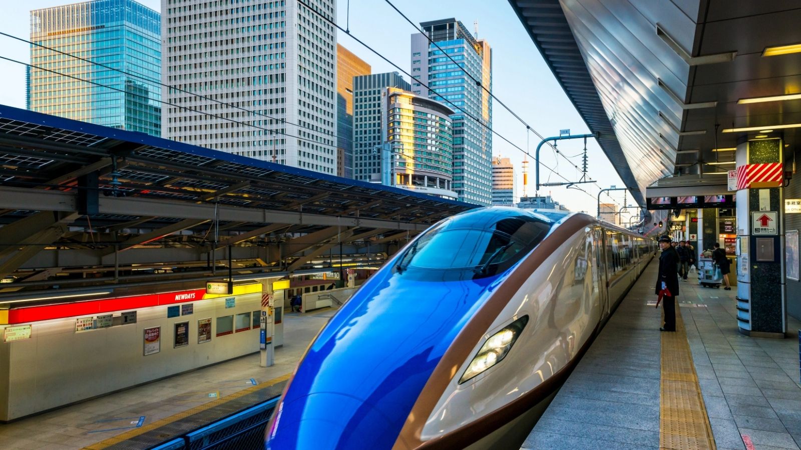 A modern bullet train arrives at an urban station with tall city buildings in the background.