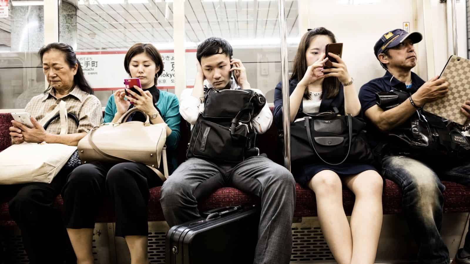 Five people sitting on a subway, all looking at their phones or listening to music, with bags on their laps.