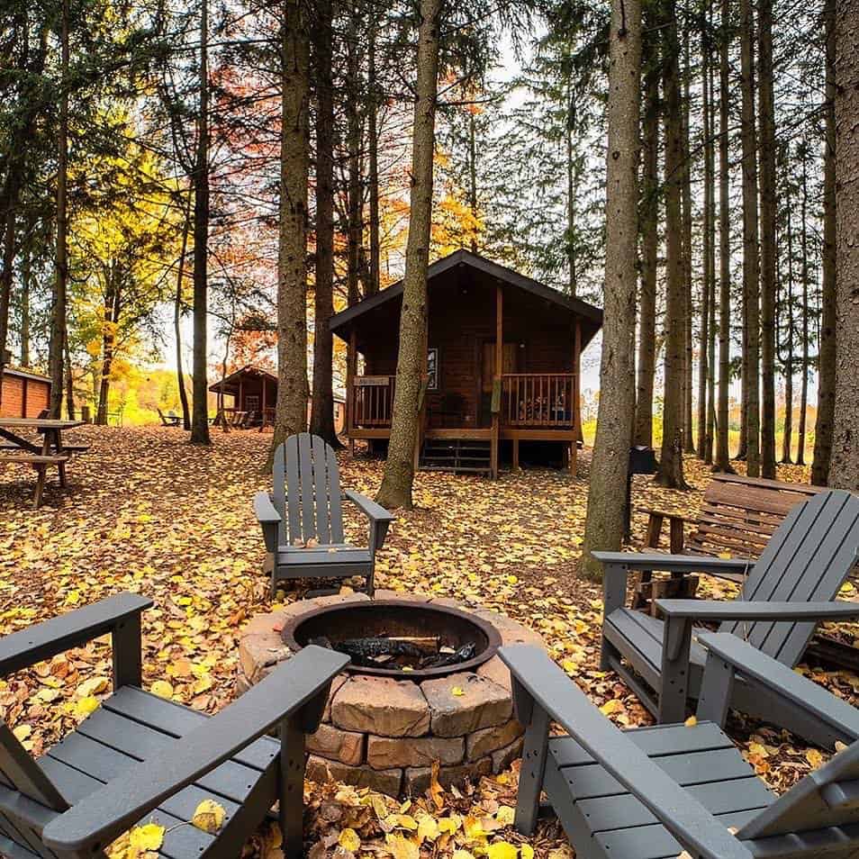 Four Adirondack chairs circle a fire pit in a forest clearing, surrounded by cabins and autumn leaves&mdash;an inviting retreat reminiscent of cozy Finger Lakes wineries with lodging.
