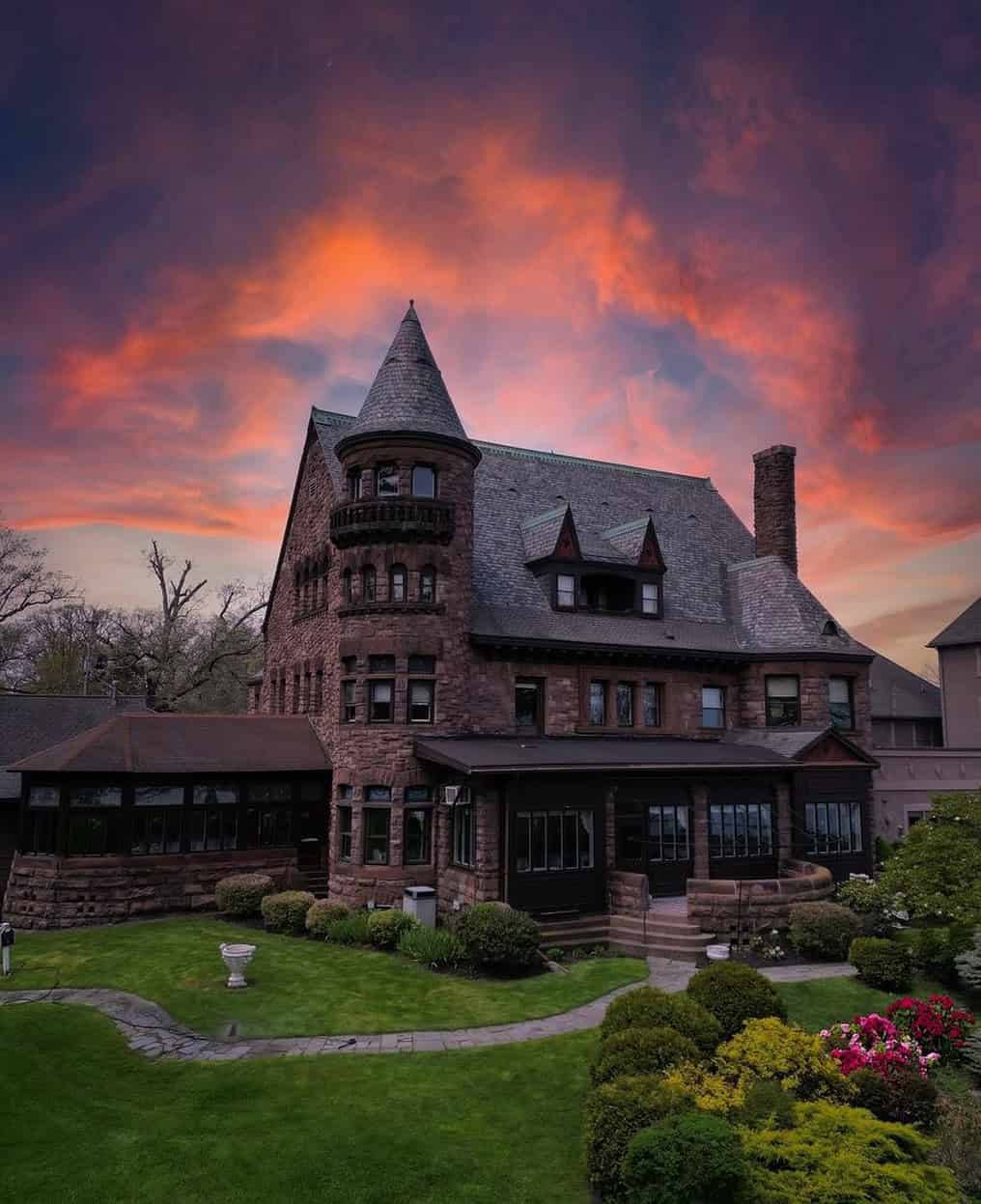 A large stone mansion with a tower under a dramatic, colorful sunset sky, surrounded by green lawns and bushes&mdash;an inviting retreat reminiscent of the elegance found at Finger Lakes Wineries with Lodging.