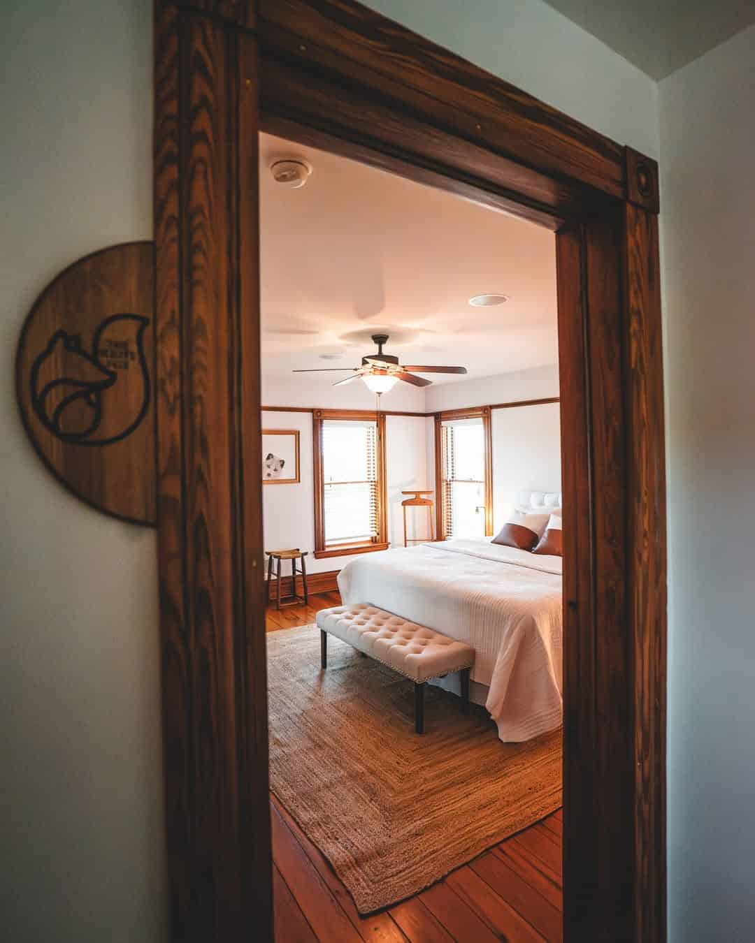View through a wooden doorway into a cozy bedroom with a white bed, bench, rug, and ceiling fan&mdash;perfect for relaxing after exploring Finger Lakes Wineries with Lodging nearby.