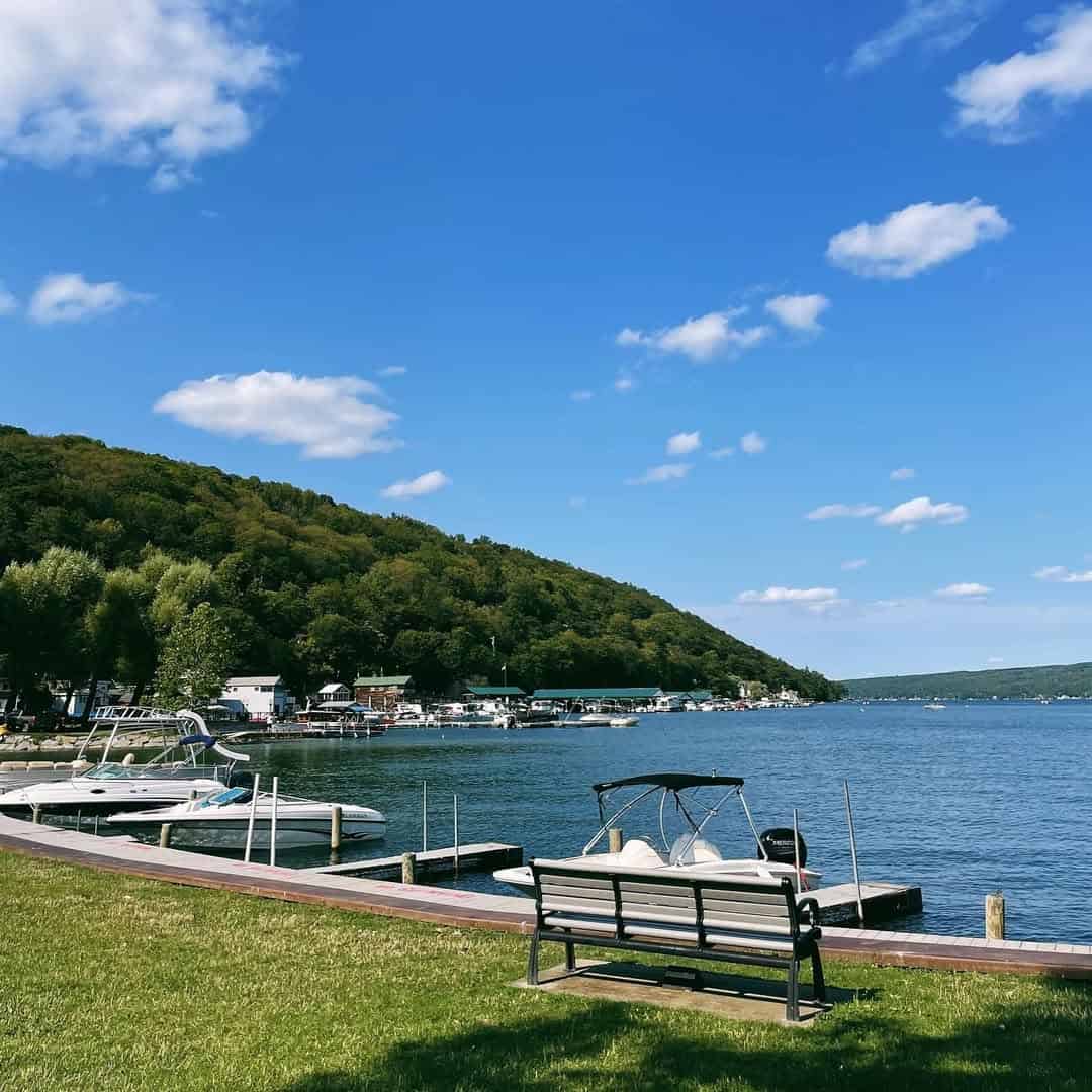 A bench overlooks a calm lake with boats and green hills under a bright blue sky with scattered clouds, offering a relaxing view near Finger Lakes Wineries with Lodging.