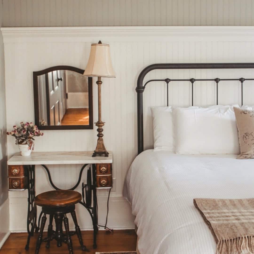 Vintage bedroom with iron bed, white bedding, an antique desk with mirror, lamp, and wooden stool&mdash;perfect for relaxing after a day exploring Finger Lakes Wineries with Lodging nearby.