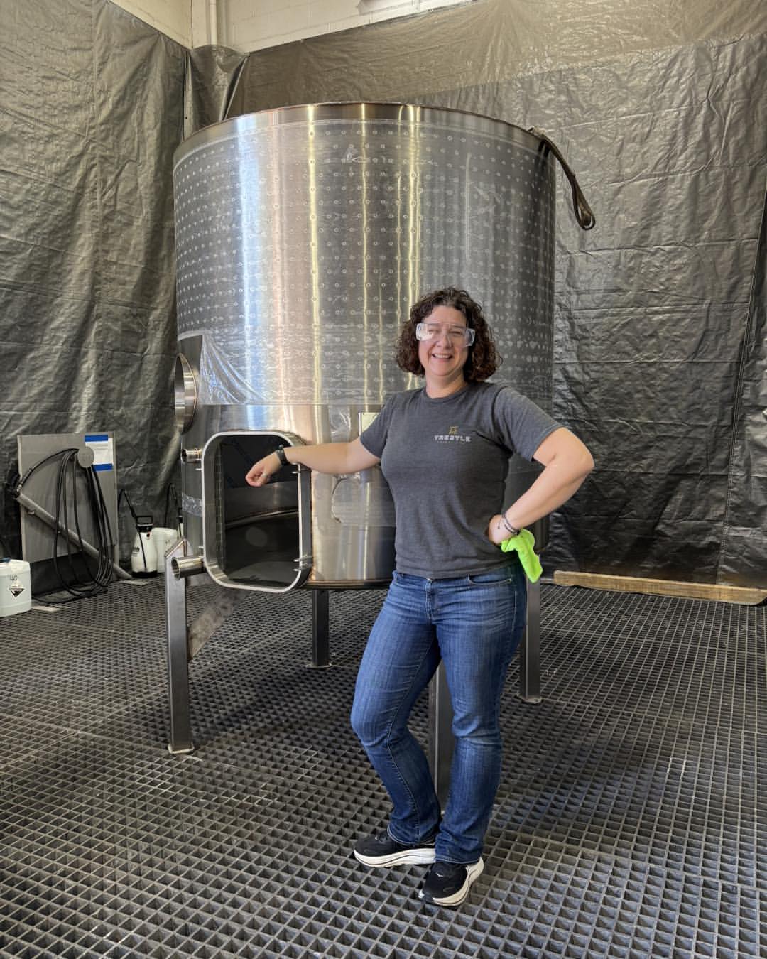 Woman in jeans and a t-shirt stands smiling by a large stainless steel fermentation tank, capturing the spirit of the best wineries in Finger Lakes in an industrial setting.