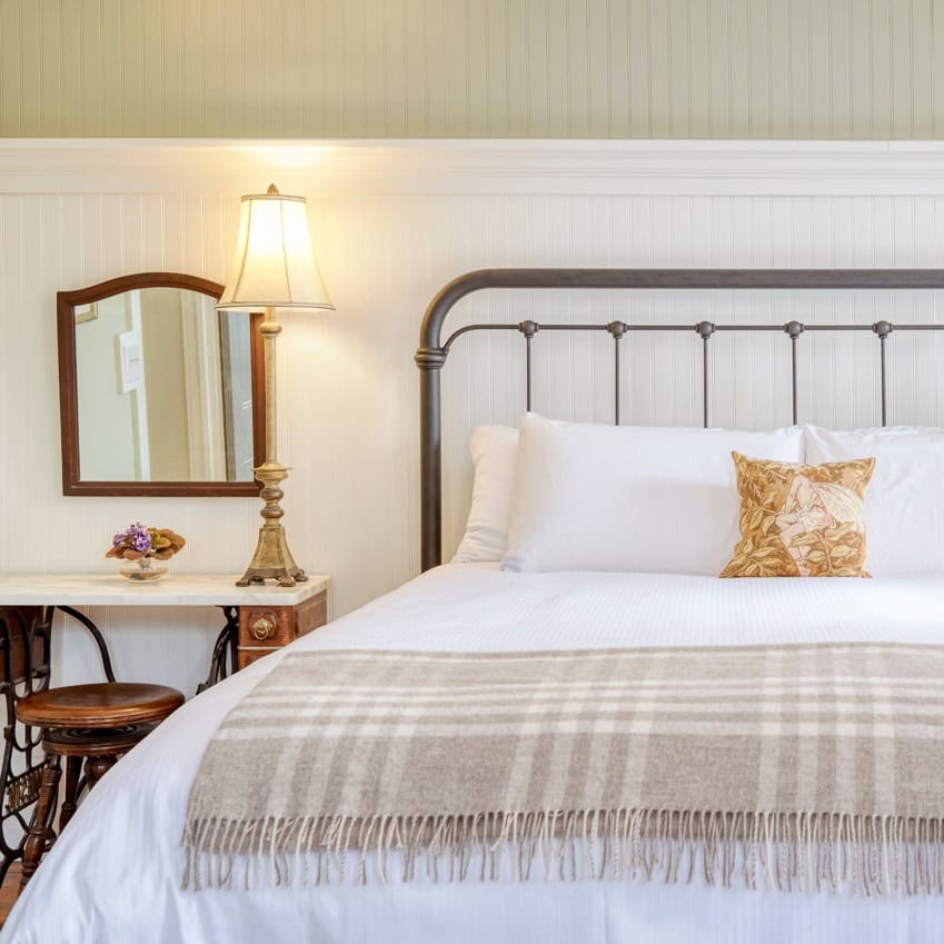 A cozy bed with white linens, a plaid blanket, and a lamp on a bedside table in a bright room&mdash;perfect for relaxing after touring Finger Lakes Wineries with Lodging.