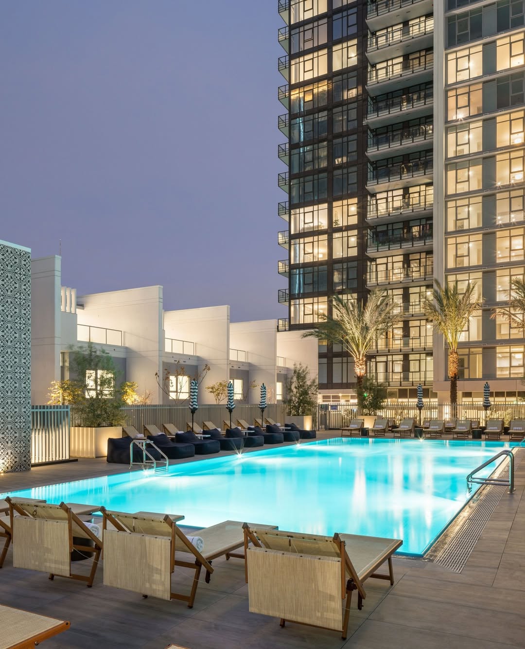 Modern apartment building with a lit outdoor pool, lounge chairs, and palm trees at dusk&mdash;reminiscent of the relaxing atmosphere found at top family resorts.
