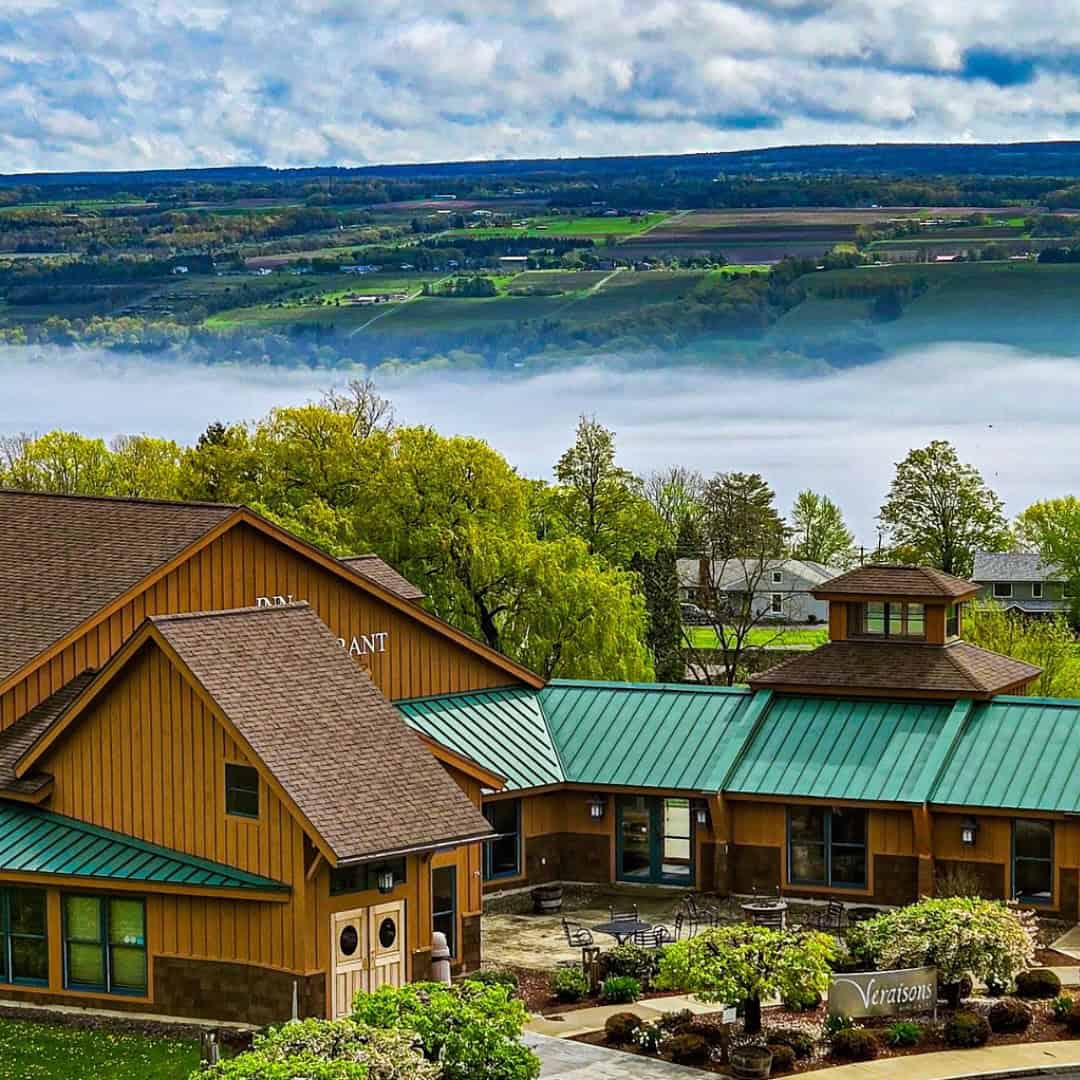 A rustic building with a green roof overlooks fields, trees, and a foggy valley under a cloudy sky&mdash;perfect for those seeking Finger Lakes wineries with lodging.