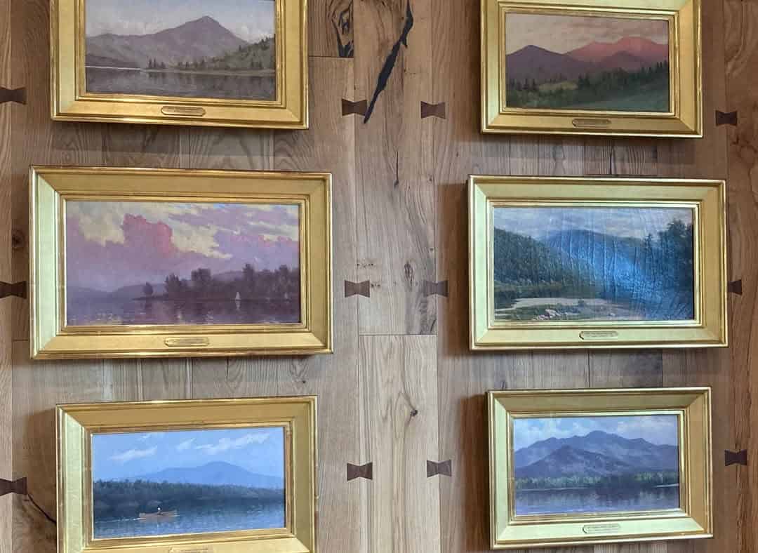 Five framed landscape paintings of mountains and lakes hang on a wooden wall with decorative inlays, evoking the tranquil ambiance of a spa at Skaneateles Fields Resort in the Finger Lakes.