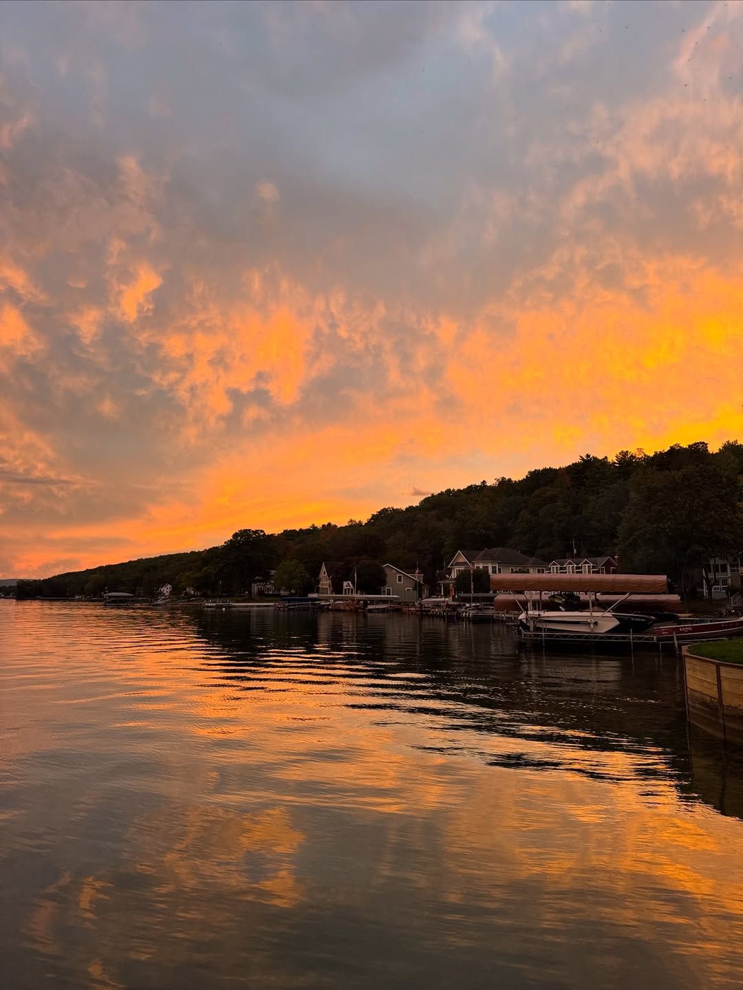Orange sunset sky reflects on calm water by a Finger Lakes lakeside, with houses, boats, and tree-covered hills—perfect for a weekend getaway.