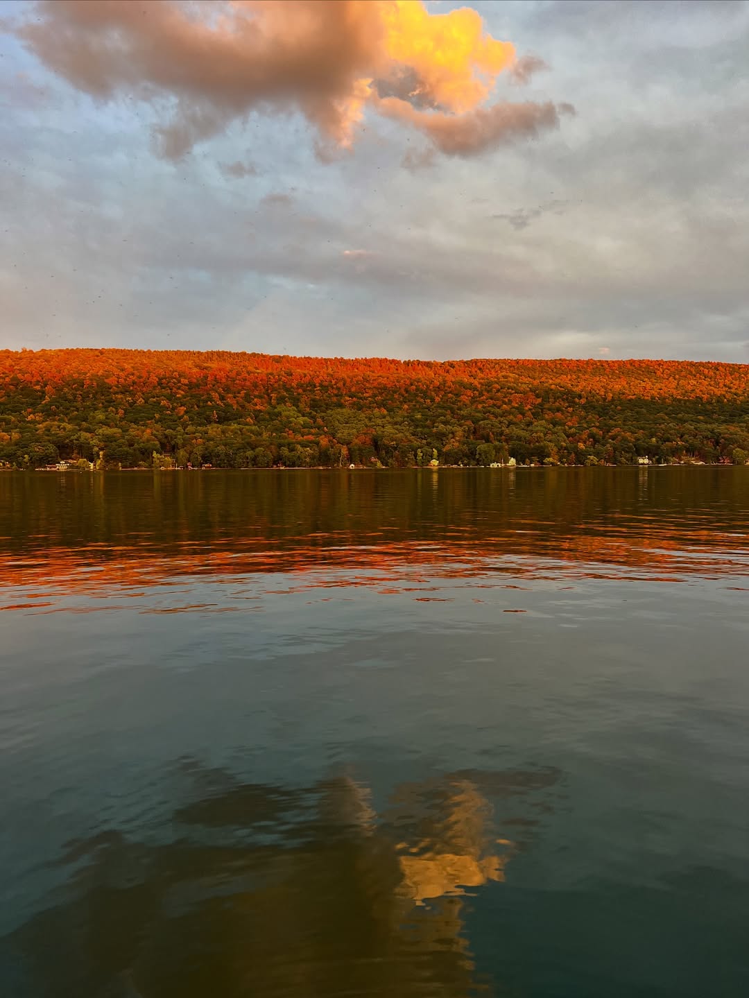 Golden sunlight hits a tree-covered hill in the Finger Lakes, reflecting on a calm lake under a partly cloudy sky—the perfect scene for a fall road trip or weekend getaway.