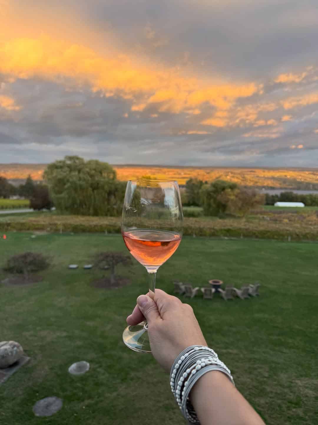 A hand holding a glass of ros&eacute; wine overlooks a green lawn at sunset with dramatic clouds, evoking the tranquil ambiance found at Finger Lakes Wineries with Lodging.