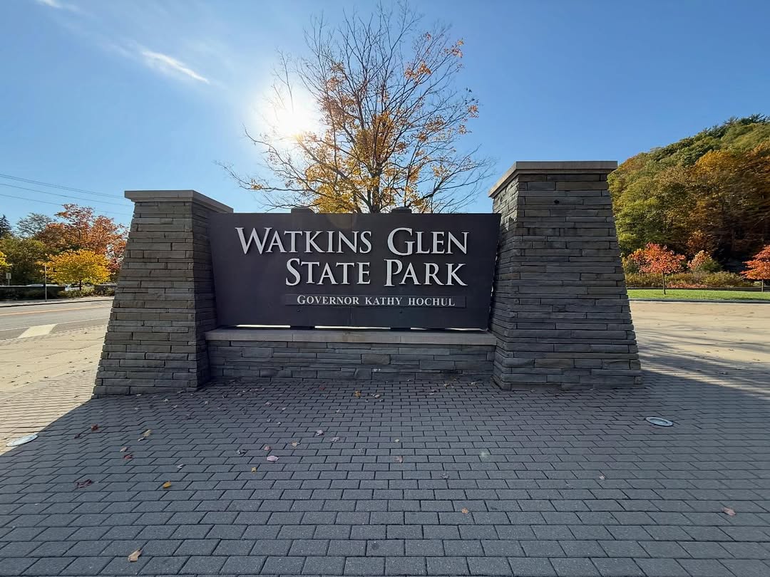 Stone sign for Watkins Glen State Park with autumn trees and sunlight in the background, welcoming visitors to the scenic trails of Watkins Glen State Park.