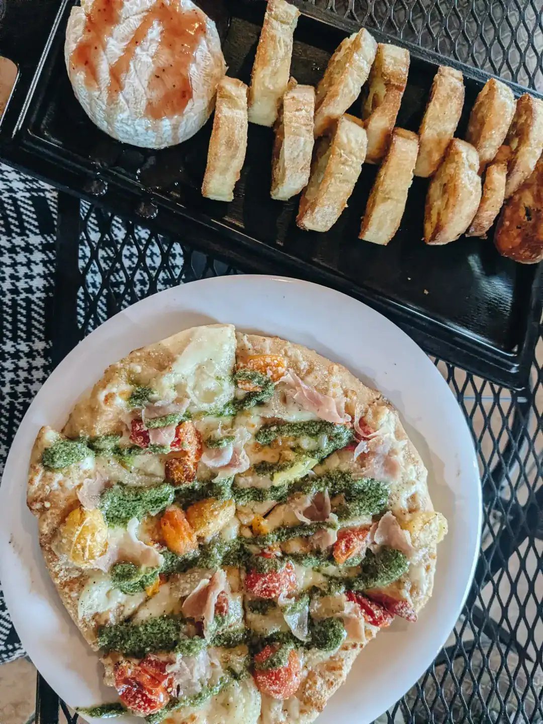 Flatbread pizza with pesto, prosciutto, and tomatoes on a plate; sliced bread and cheese on a tray above&mdash;perfect bites to enjoy after touring Finger Lakes Wineries with Lodging.