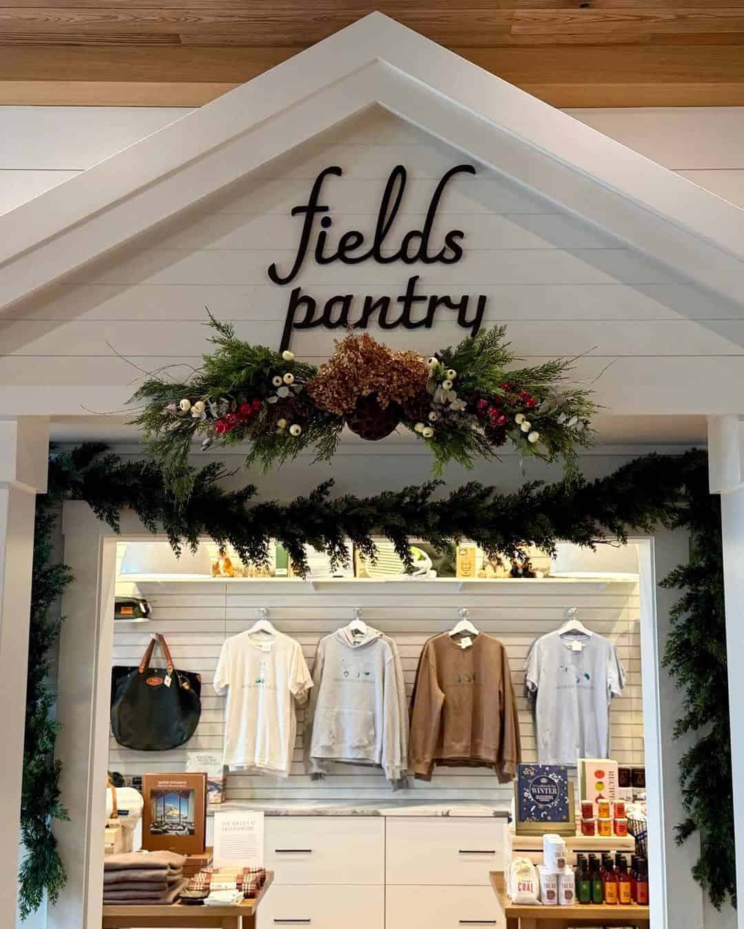 Gift shop entrance at Skaneateles Fields Resort & Spa, with "fields pantry" sign, floral garland, and a variety of Finger Lakes-inspired clothing and goods displayed inside.