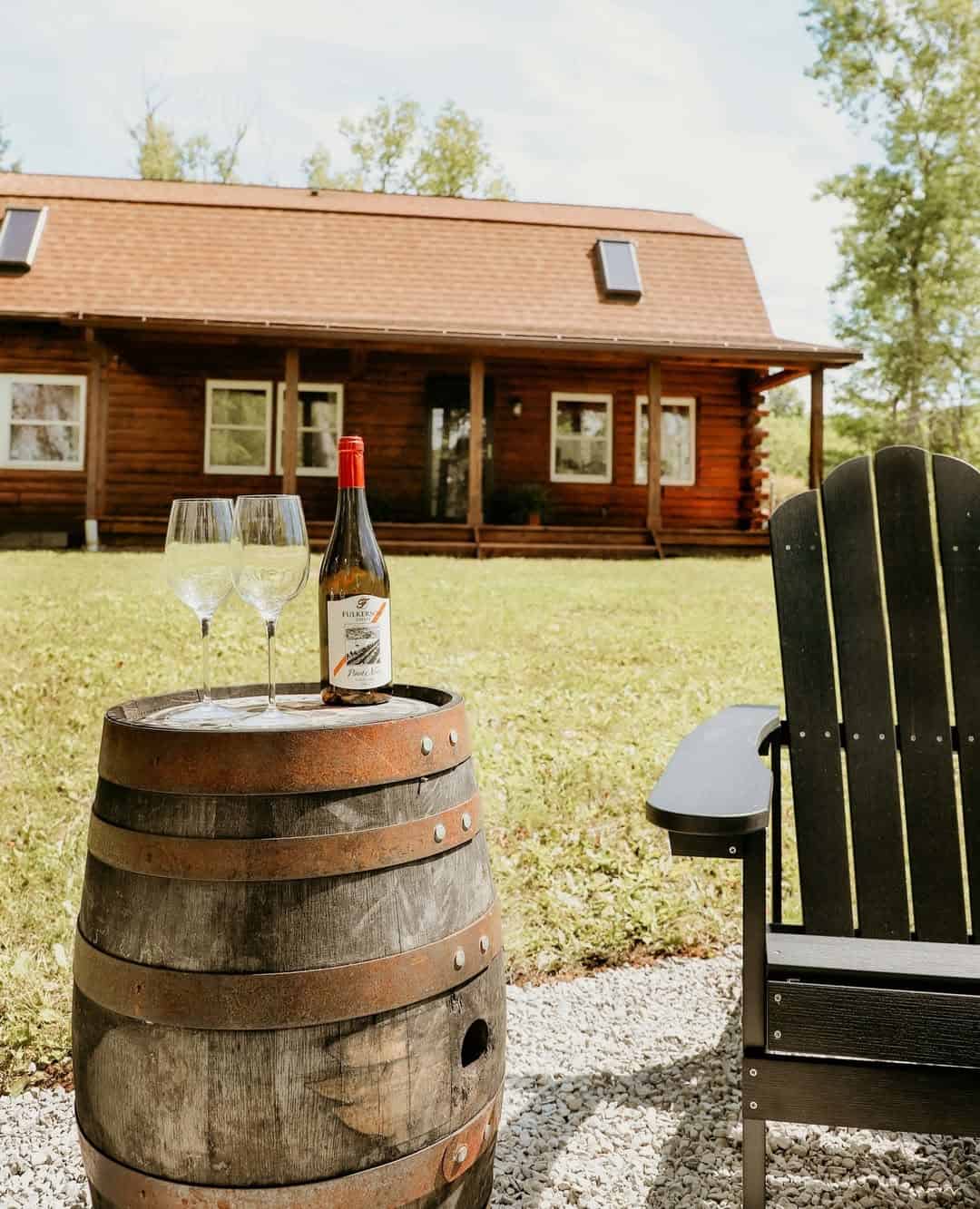 Wine bottle and glasses on a barrel beside a chair, with a log cabin in the background on a sunny day&mdash;perfect for relaxing at one of the Finger Lakes wineries with lodging.