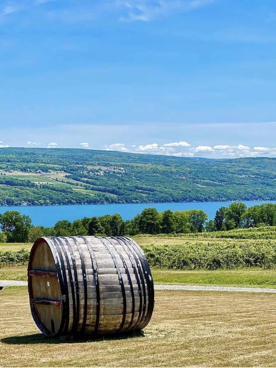 A large wooden barrel on grass with a lake and green hills in the background under a blue sky, capturing the scenic charm of Finger Lakes wineries with lodging.