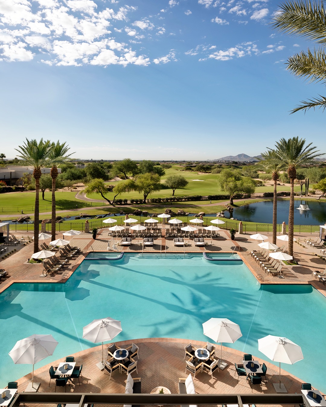 Large resort pool with lounge chairs, umbrellas, palm trees, and a golf course in the background under a blue sky&mdash;one of the best family resorts for a memorable getaway.