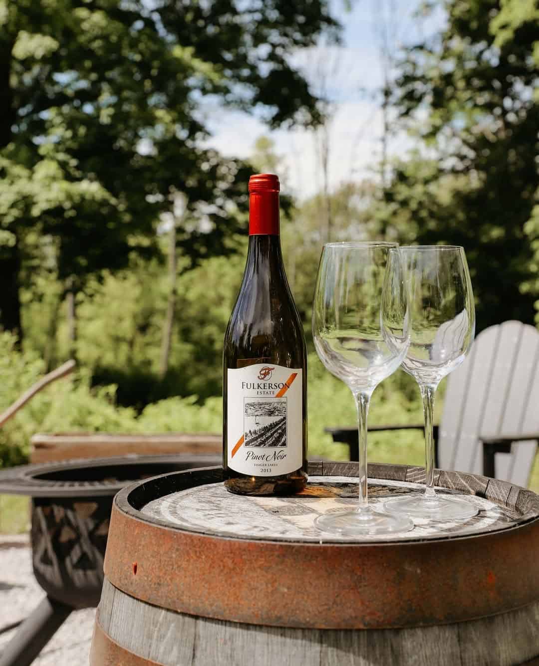 A wine bottle and two empty glasses rest on a barrel outdoors at one of the Finger Lakes Wineries with Lodging, surrounded by trees and inviting chairs in the background.
