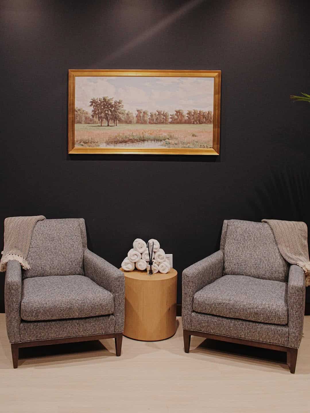 Two gray armchairs with blankets, a round wooden table, and a landscape painting on a dark wall create a cozy retreat reminiscent of Finger Lakes charm, perfect for unwinding after a day at the Skaneateles Fields Resort & Spa.