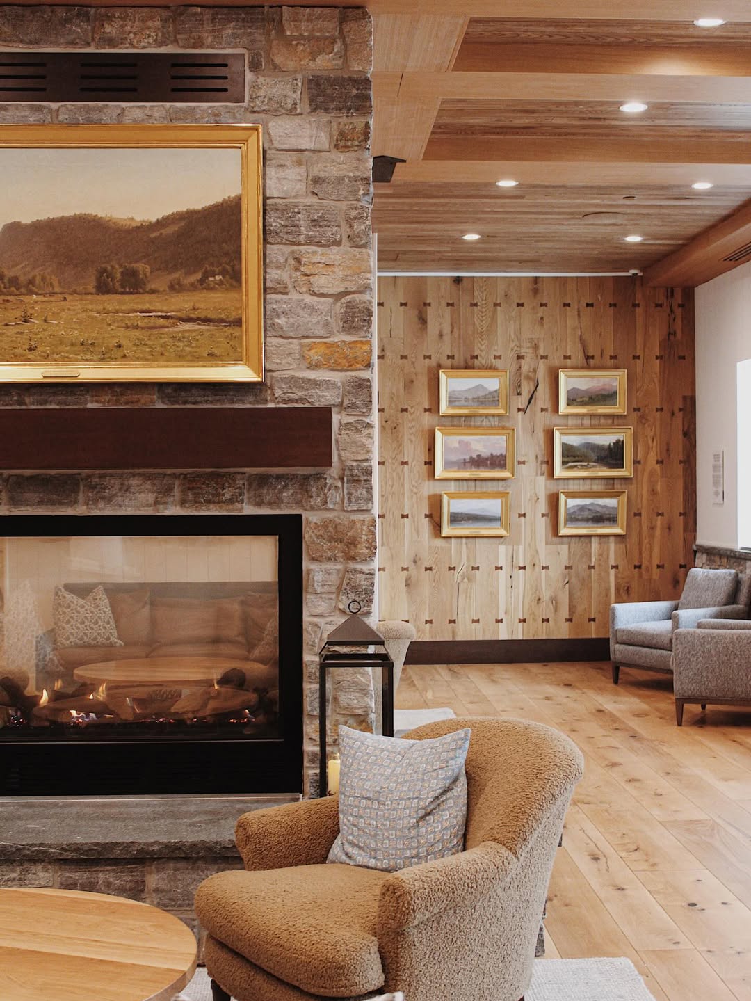 Cozy living room with fireplace, armchairs, wood paneling, and landscape paintings on the walls—perfect for unwinding after a day at Skaneateles Fields Resort & Spa in the beautiful Finger Lakes.
