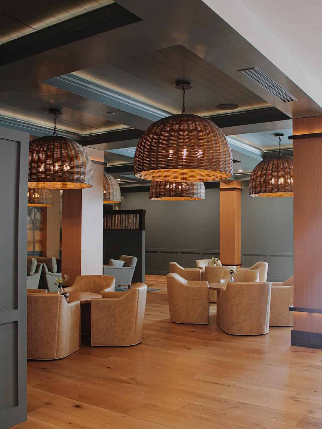 A modern lounge at Skaneateles Fields Resort features tan armchairs, round tables, and large woven pendant lights on a wooden floor—perfect for relaxing after a day at the spa in the scenic Finger Lakes.