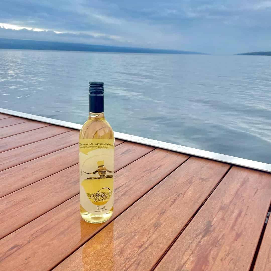 A bottle of white wine on a wooden dock overlooking a calm lake under a cloudy sky, evoking the tranquil charm of Finger Lakes wineries with lodging.