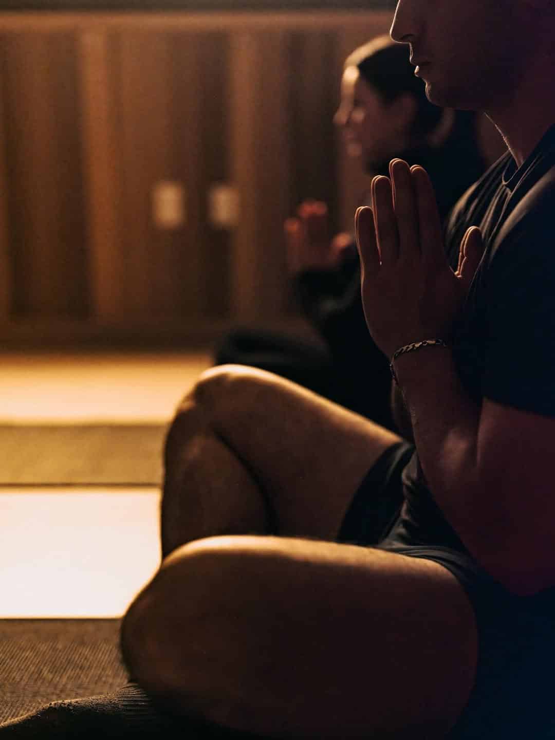 Two people sit cross-legged on yoga mats with hands in prayer position, meditating in soft indoor light at Skaneateles Fields Resort, a tranquil spa retreat in the heart of Finger Lakes.