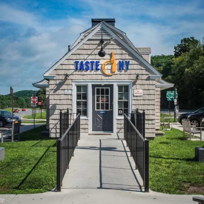 Small gray roadside shop with "Taste NY" sign, ramp entrance, and green trees in the background—perfect for a quick stop on your Fall Road Trip or Weekend Getaway through the Finger Lakes.