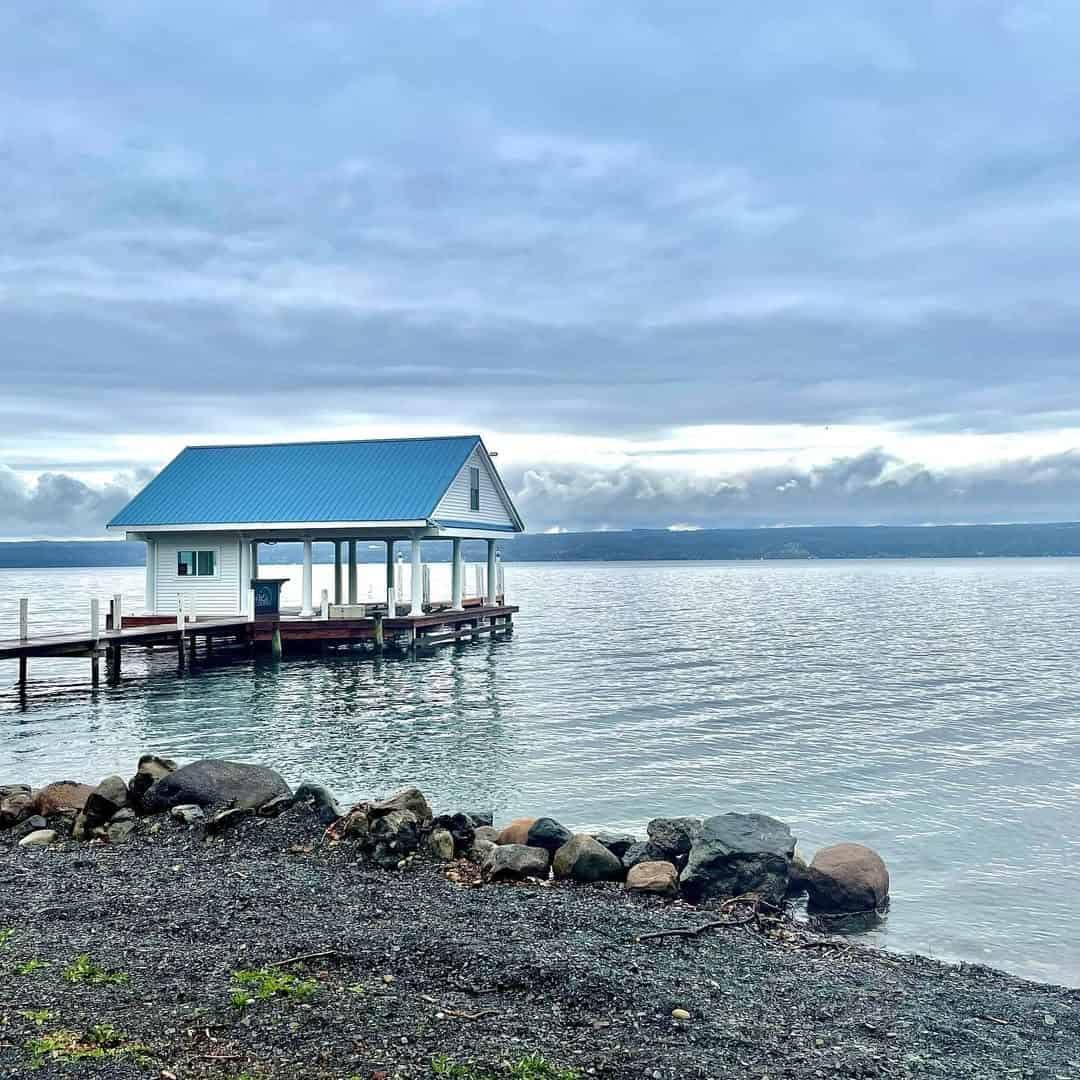 Small boathouse with a blue roof on a calm lake near Finger Lakes wineries with lodging, cloudy sky above, and rocky shoreline in the foreground.