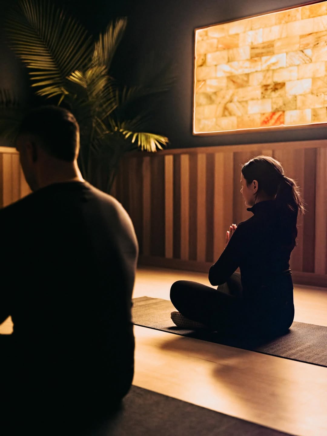 Two people in dark clothing sit cross-legged on yoga mats in a calm, softly lit room with a glowing wall panel at Skaneateles Fields Resort & Spa, nestled in the tranquil beauty of the Finger Lakes.