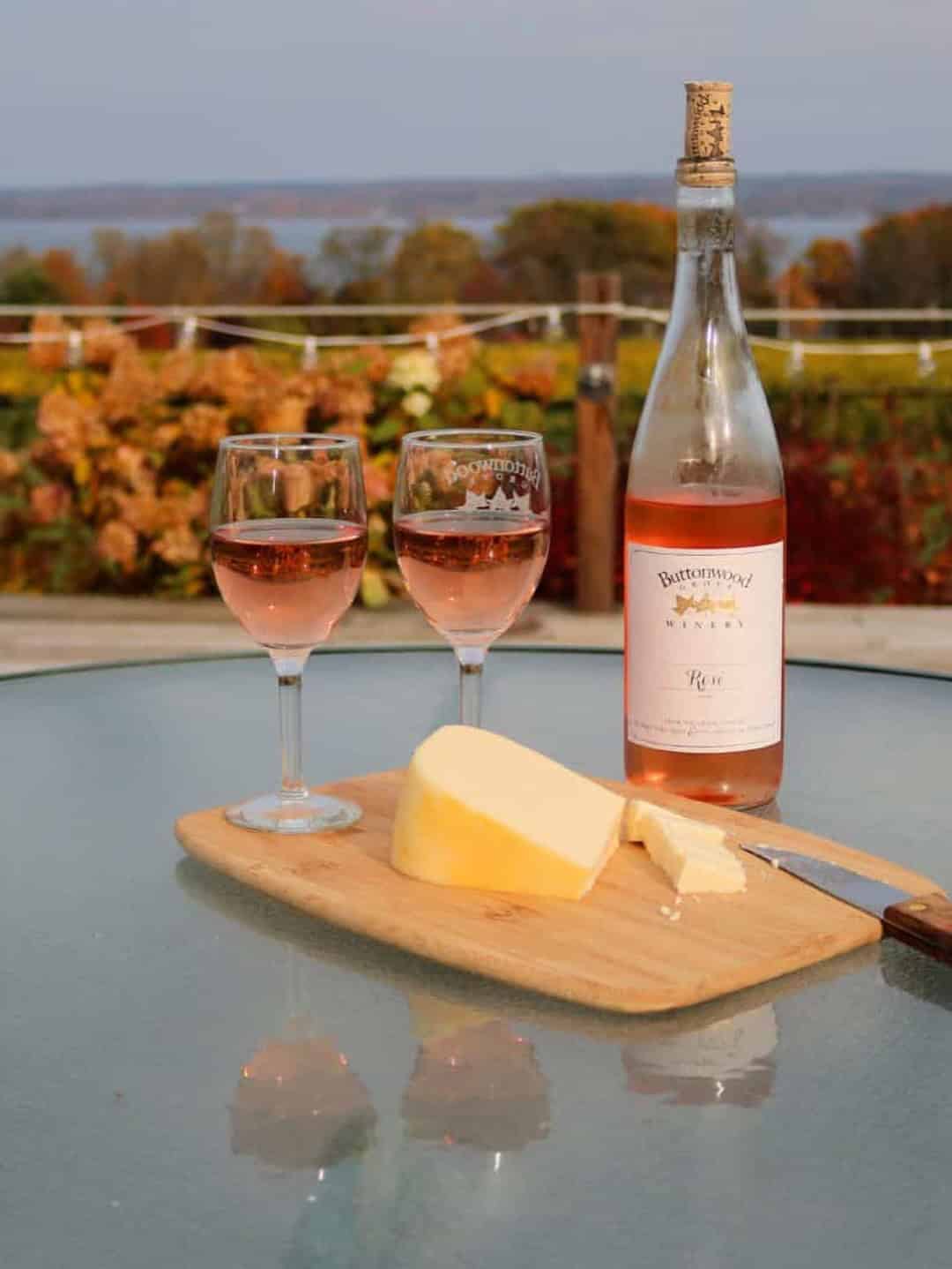 Two glasses and a bottle of ros&eacute; with cheese on a board, set on an outdoor table overlooking vineyards&mdash;experience moments like this at Finger Lakes Wineries with Lodging.