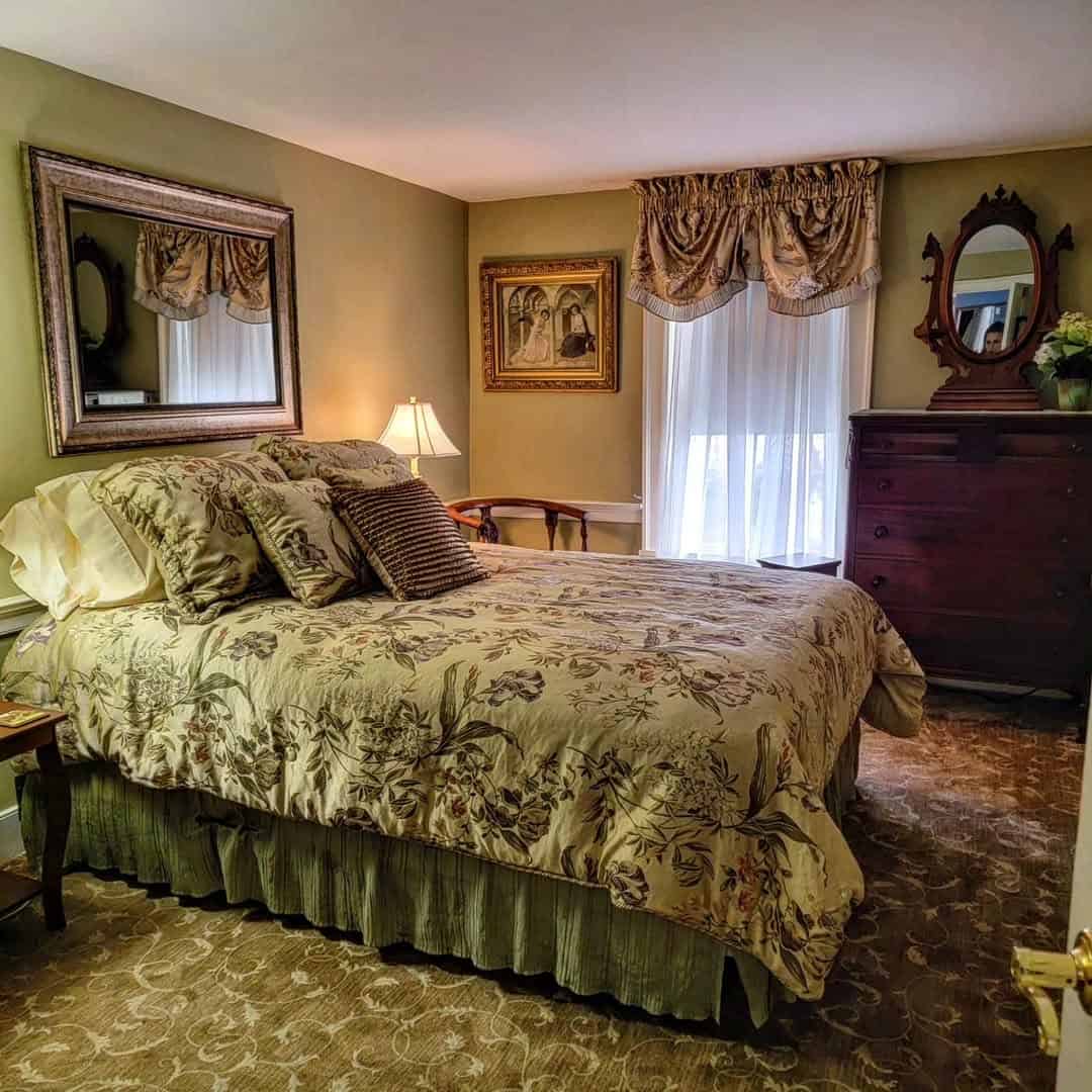 Cozy bedroom with a floral bedspread, wooden furniture, framed art, and soft natural light through curtained windows&mdash;perfect for relaxing after a day exploring Finger Lakes Wineries with Lodging nearby.