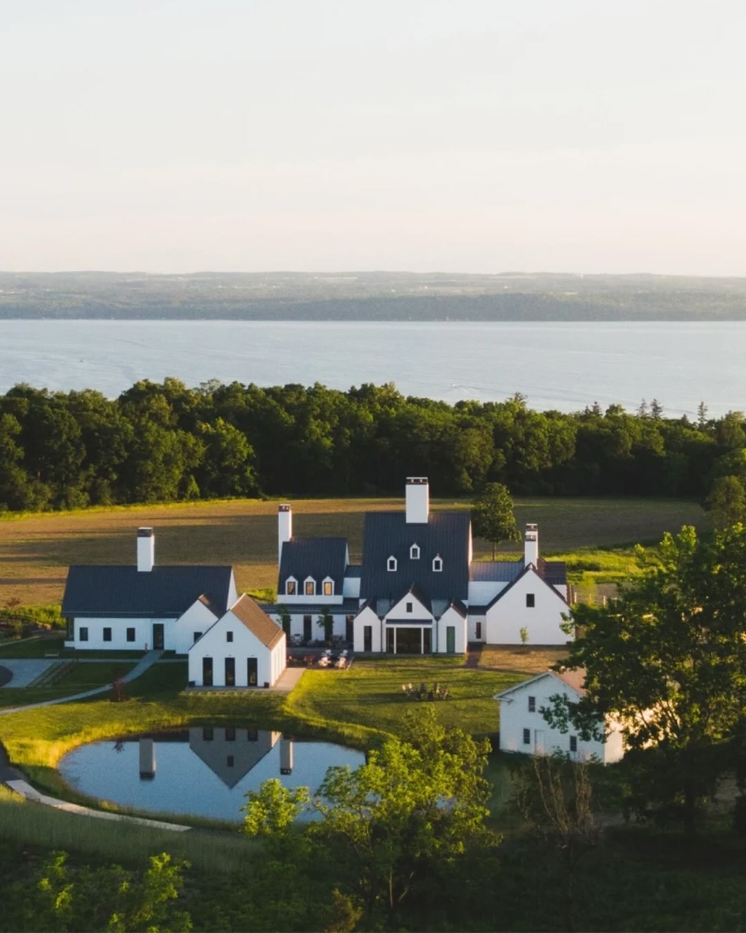 Aerial view of a modern white farmhouse with a pond, surrounded by trees and fields near a large body of water—perfect for a Finger Lakes weekend getaway.