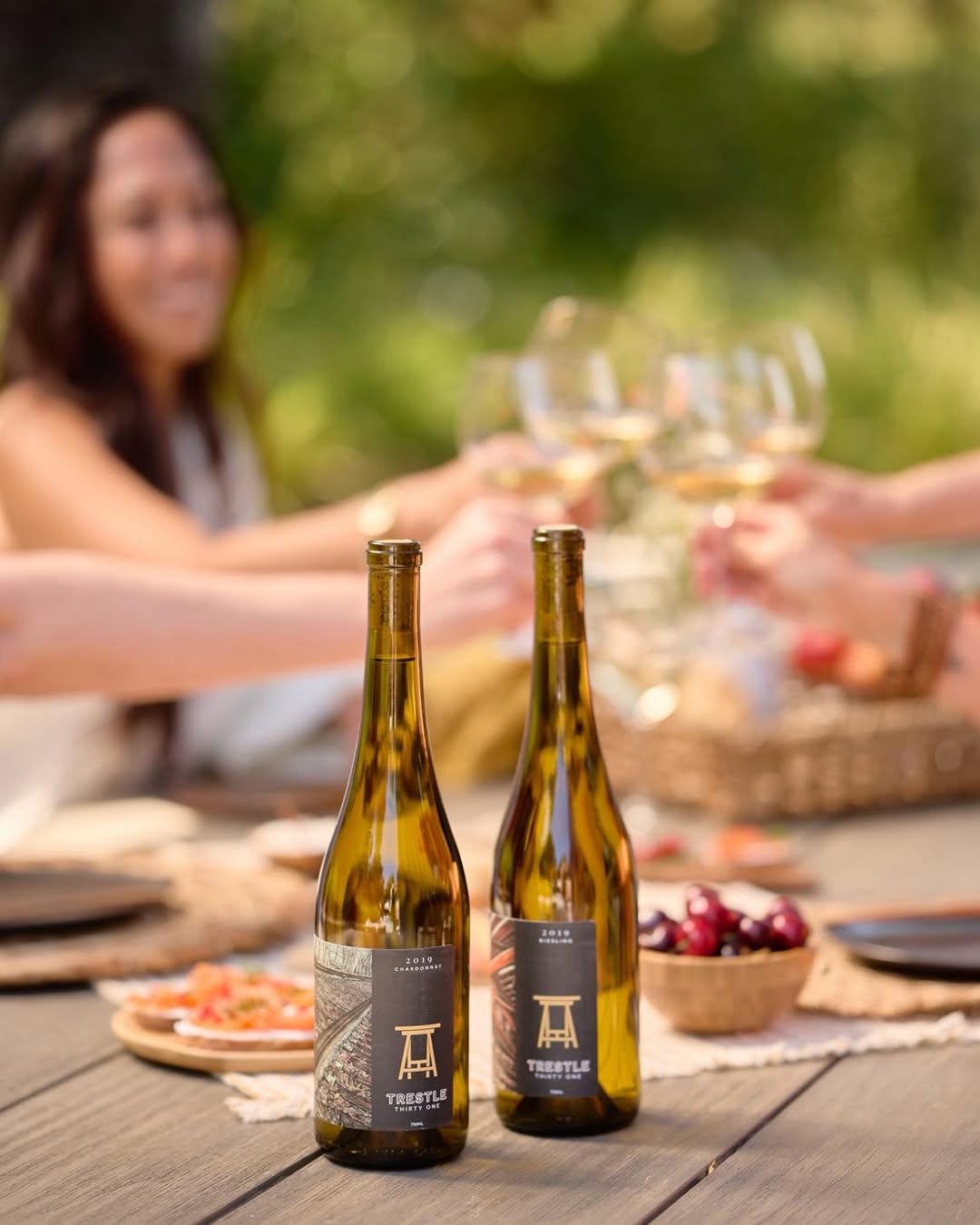 Two wine bottles from the best wineries in Finger Lakes sit on an outdoor table, with friends toasting in the background and delicious food dishes nearby.