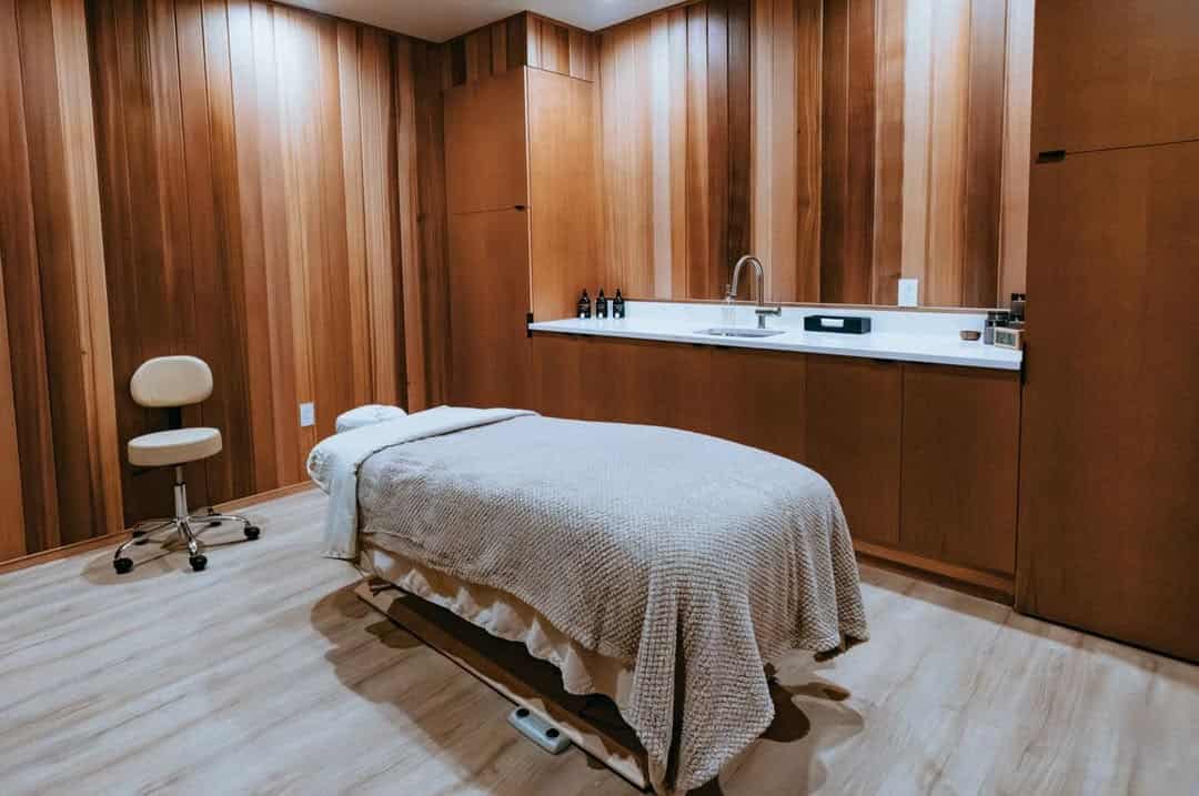 A massage table with a beige blanket in a wooden spa room at Skaneateles Fields Resort, with a sink and stool nearby, offering relaxation in the heart of the Finger Lakes.