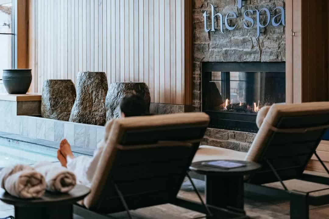 Unwind on a lounge chair by the fireplace at the luxurious Spa in Skaneateles Fields Resort, surrounded by elegant stone décor and plush towels—an idyllic Finger Lakes escape.