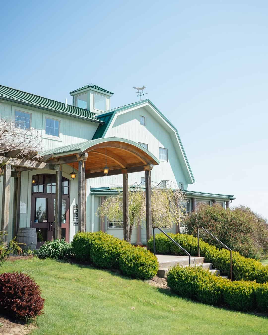 Large light-green barn building with a wooden entrance canopy, surrounded by neatly trimmed bushes and lawn&mdash;perfect for guests seeking Finger Lakes Wineries with Lodging.