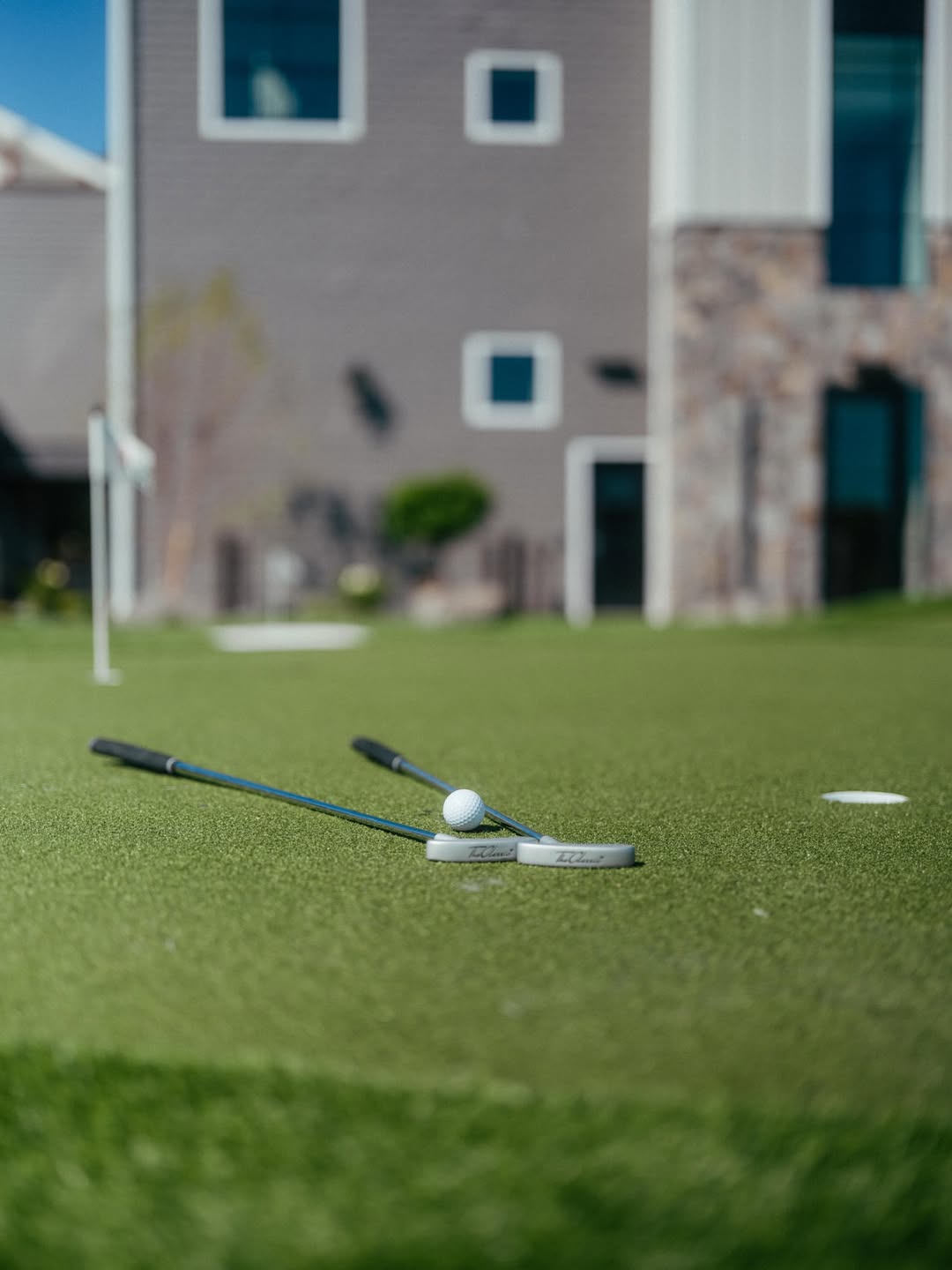 Two golf putters and a golf ball lie on a putting green at Skaneateles Fields Resort, with a building in the background, offering a perfect glimpse of recreation in the beautiful Finger Lakes region.