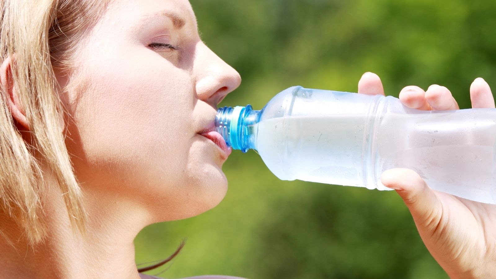 A person drinks water from a plastic bottle outdoors on a sunny day, with greenery in the background—one of the essential hot weather travel tips to stay refreshed during your adventures.