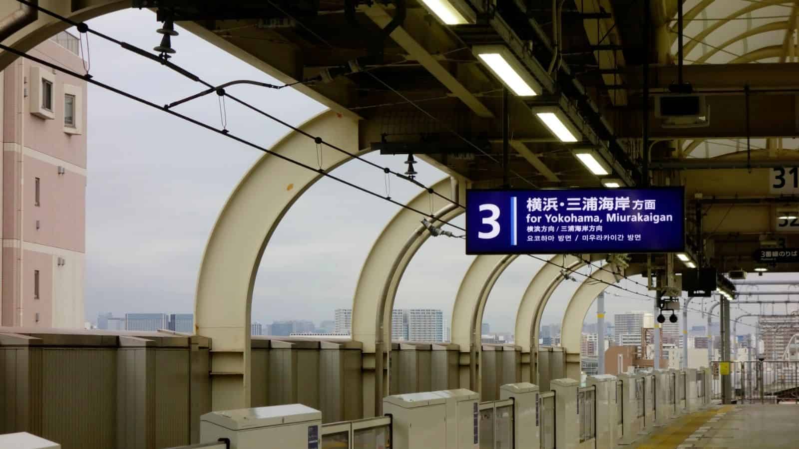 A train station platform with a sign for Yokohama and Miurakaigan, overlooking city buildings.