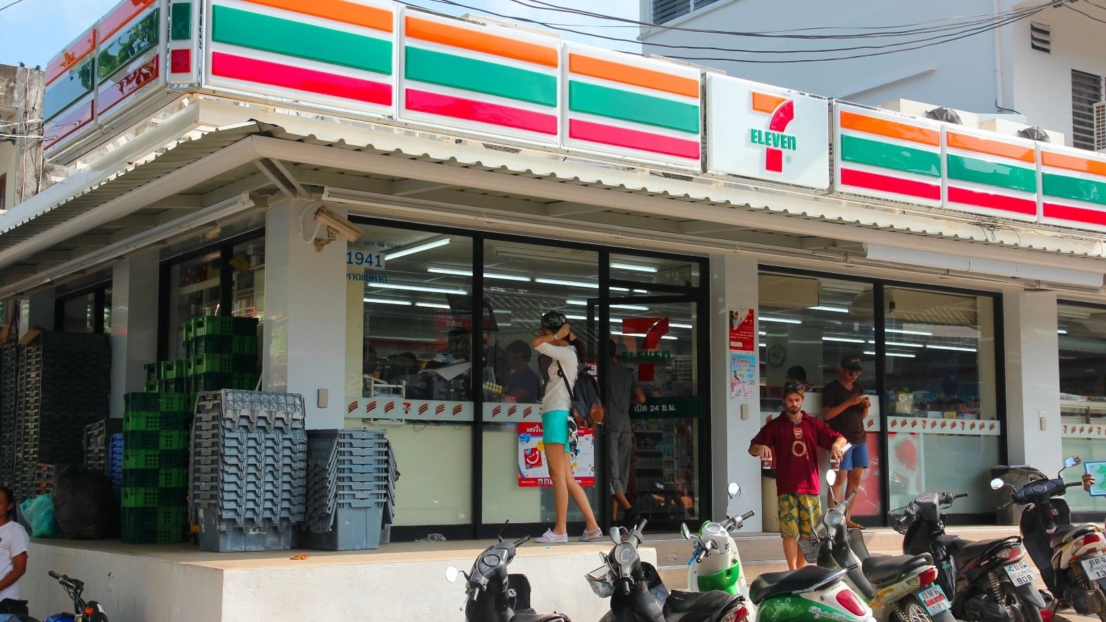 People enter and exit a 7-Eleven store with motorbikes parked outside on a sunny day—a familiar scene for those seeking Asia travel tips and hot weather travel hacks.