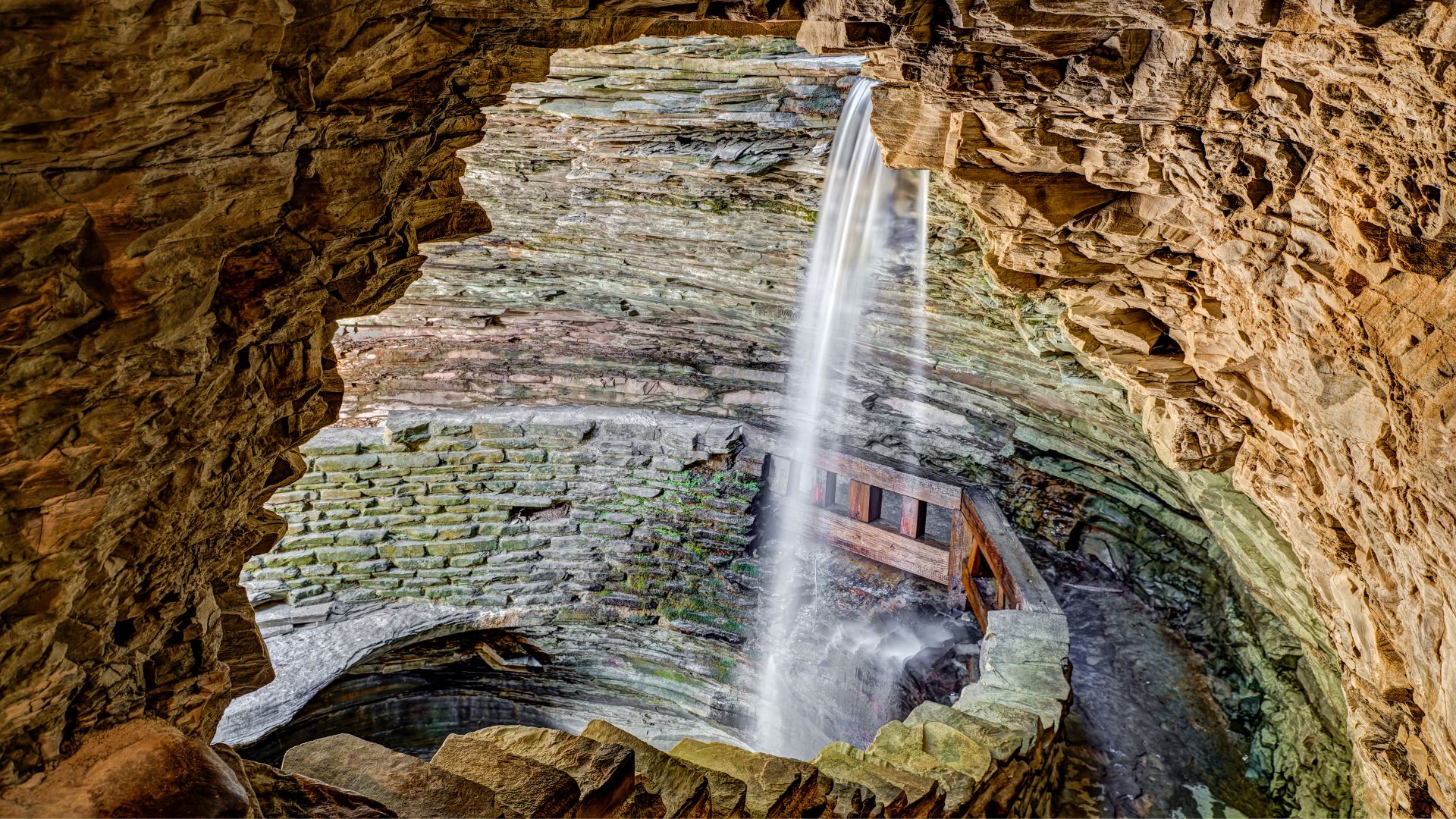 View through a rocky cave opening at Watkins Glen State Park, showcasing a waterfall flowing into a stone-walled gorge with a wooden bridge.