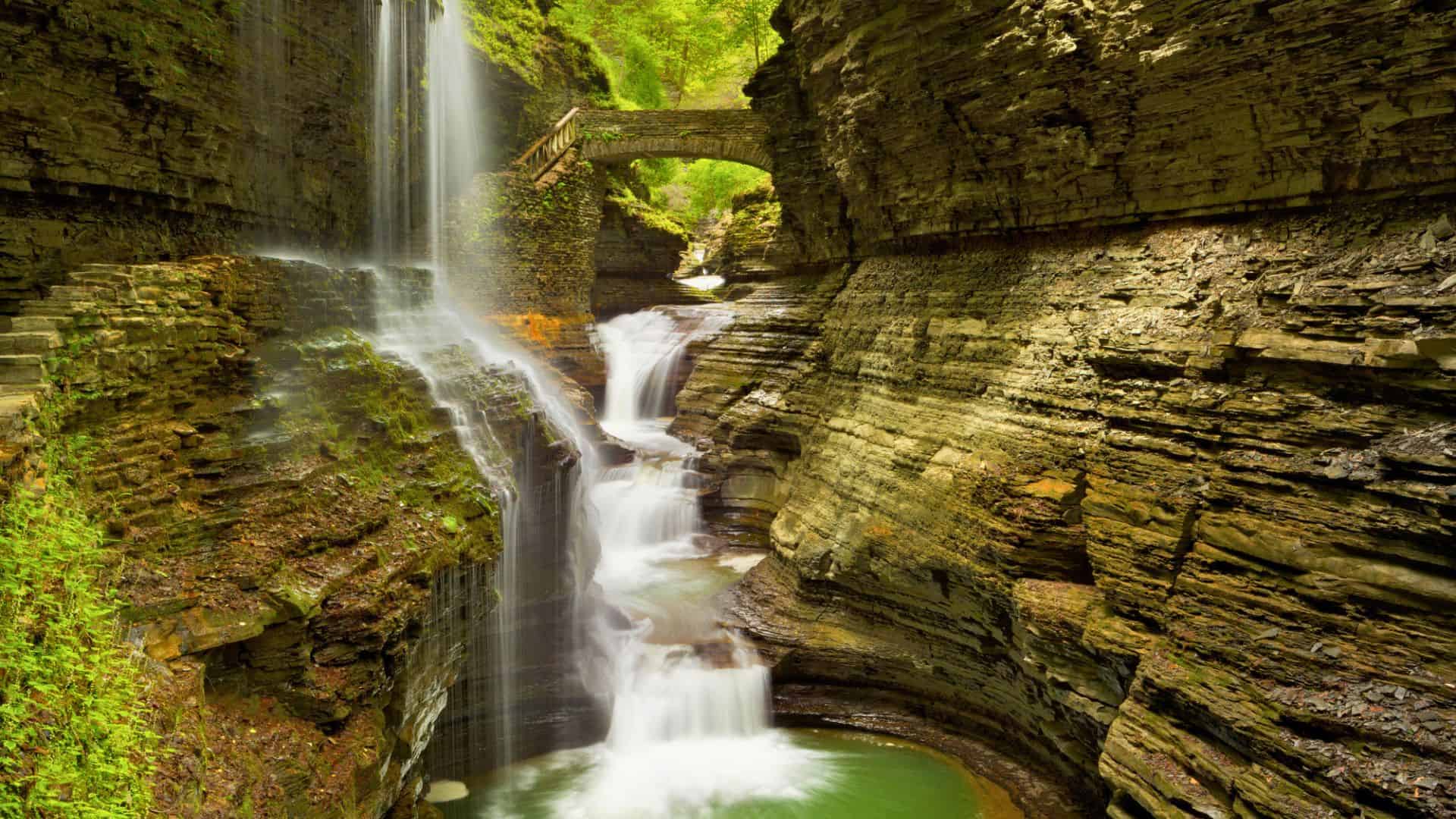 A waterfall flows through layered rock formations below a stone bridge in the lush, green gorge of Watkins Glen State Park&mdash;an ideal spot for a Family Day adventure.