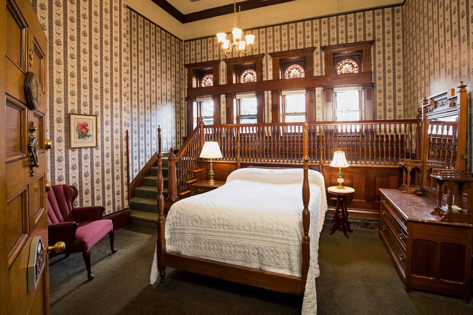 A vintage bedroom with a wood bed, white quilt, floral wallpaper, and stained glass windows above a raised area&mdash;perfect for relaxing after touring Finger Lakes Wineries with Lodging.