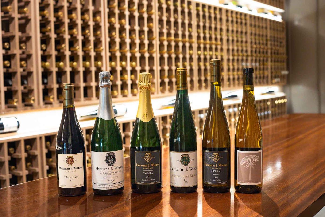Six wine bottles displayed on a wooden counter with a large wine rack filled with bottles in the background, evoking the charm of the best wineries in Finger Lakes.